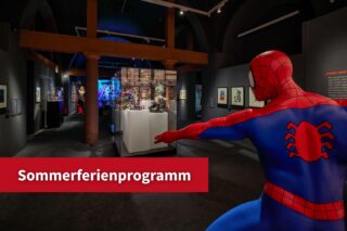 Blick in die Ausstellung "Superheroes". In der linken Ecke befindet sich ein roter Störer mit dem Hinweis "Sommerferienprogramm".