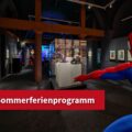 Blick in die Ausstellung "Superheroes". In der linken Ecke befindet sich ein roter Störer mit dem Hinweis "Sommerferienprogramm".