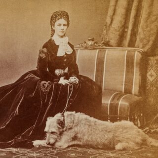 Historische Fotografie: Sisi sitzt auf einem Sofa in einem dunklen Samtkleid, vor ihr liegt ein großer heller Hund.