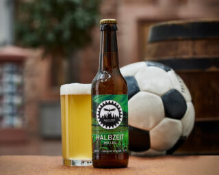 Foto des Bieres "Halbzeit-Helles" mit einem Bierglas, Fußball und Bierfass im Hintergrund.